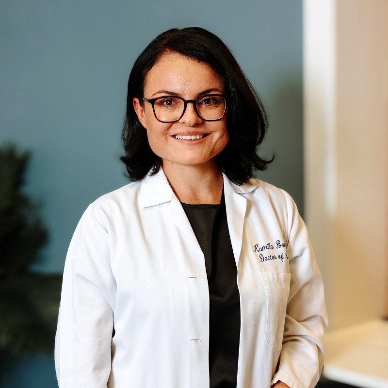 Dr. Kamila Bome, audiologist, smiling in dark-rimmed glasses and a white coat with her name stitched on it